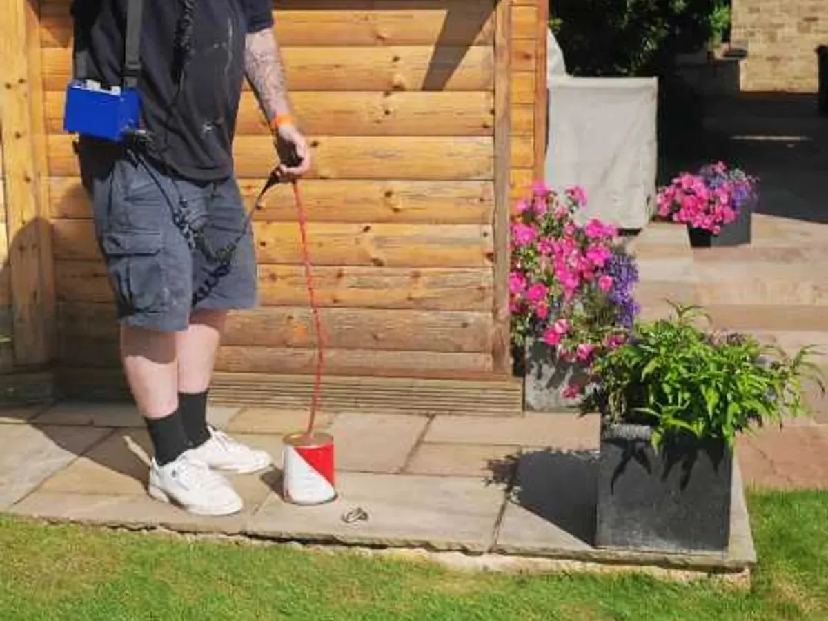 An ADI engineer with the amplifier on a shoulder strap holds a red and white ground microphone on its lead over a patio beside a timber summer house